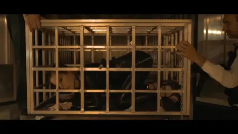 Slim brunette in ponytail watches two guys caged in dim jail setting